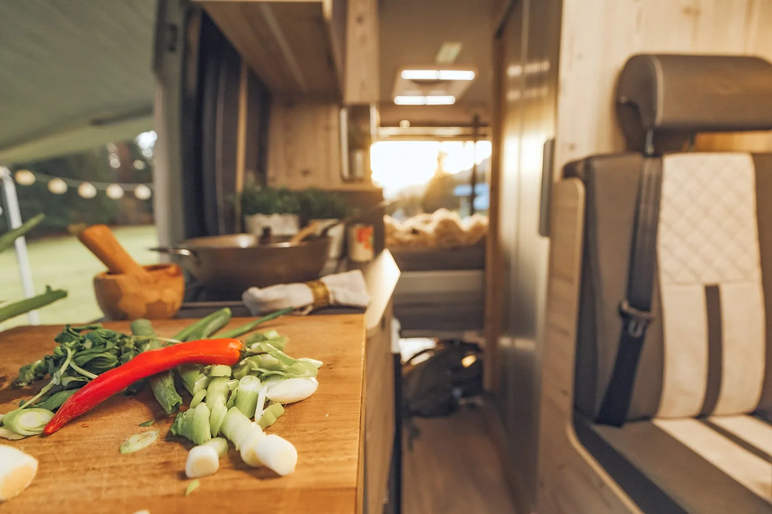 Chopped green onions and a red chili pepper on a wooden cutting board inside a camper van kitchen with a seat and bed in the background.