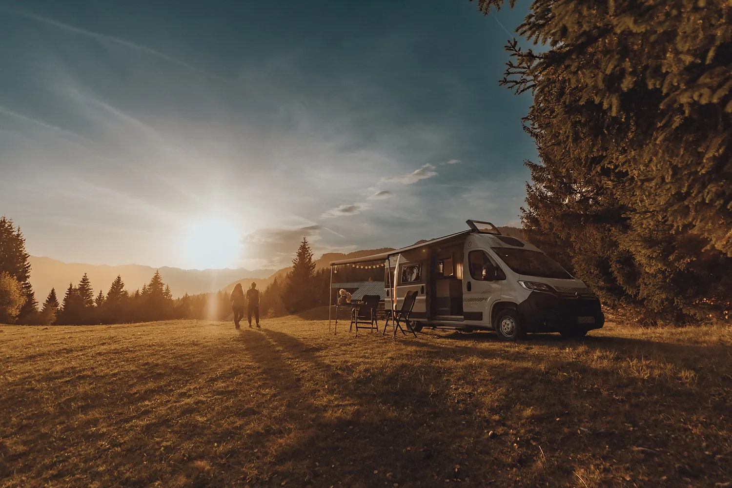 Two people walking towards the sunset near a camper van parked on a grassy field surrounded by trees.