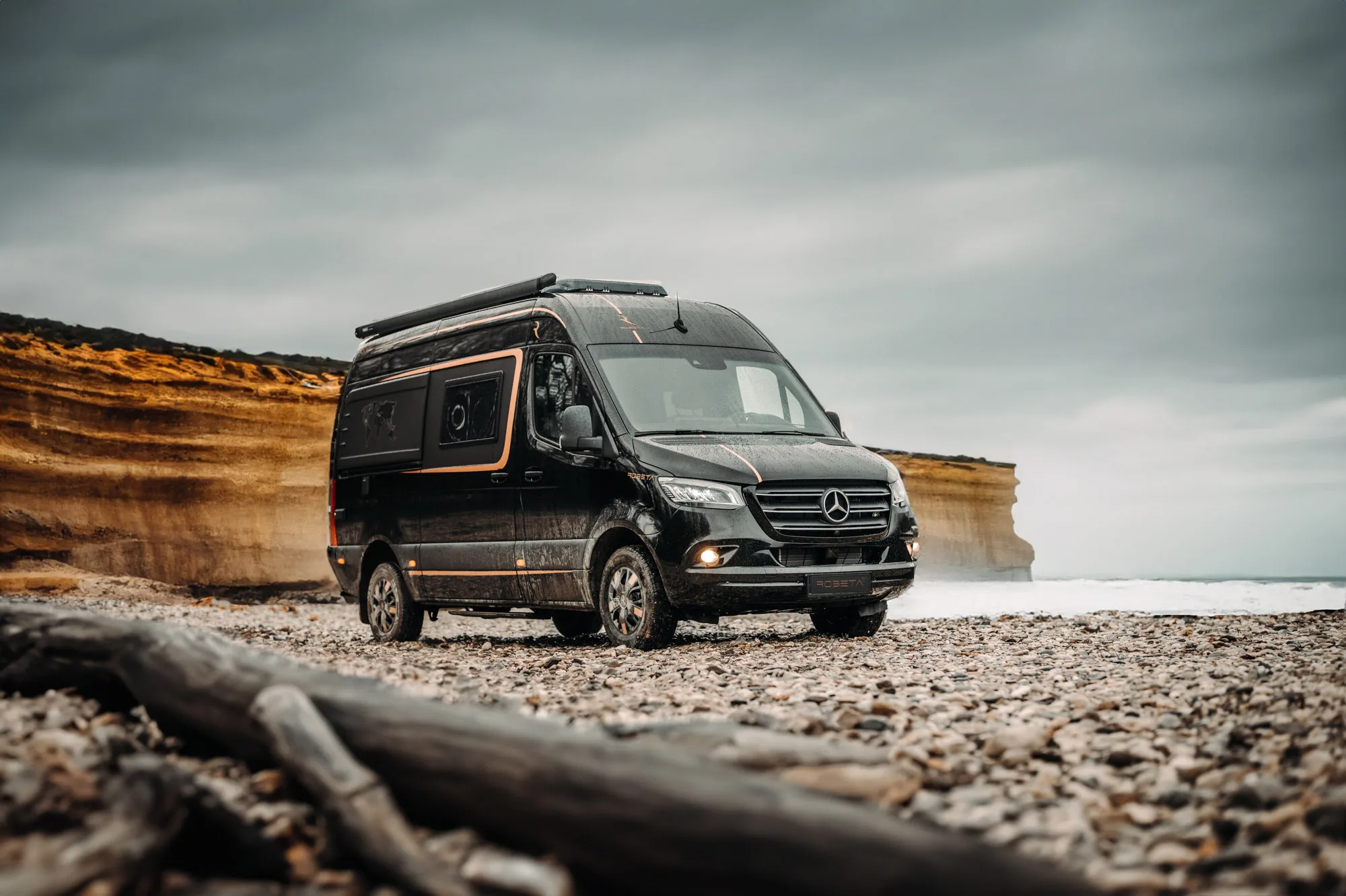 Black Mercedes camper van parked on a rocky beach with cliffs and cloudy sky in the background.