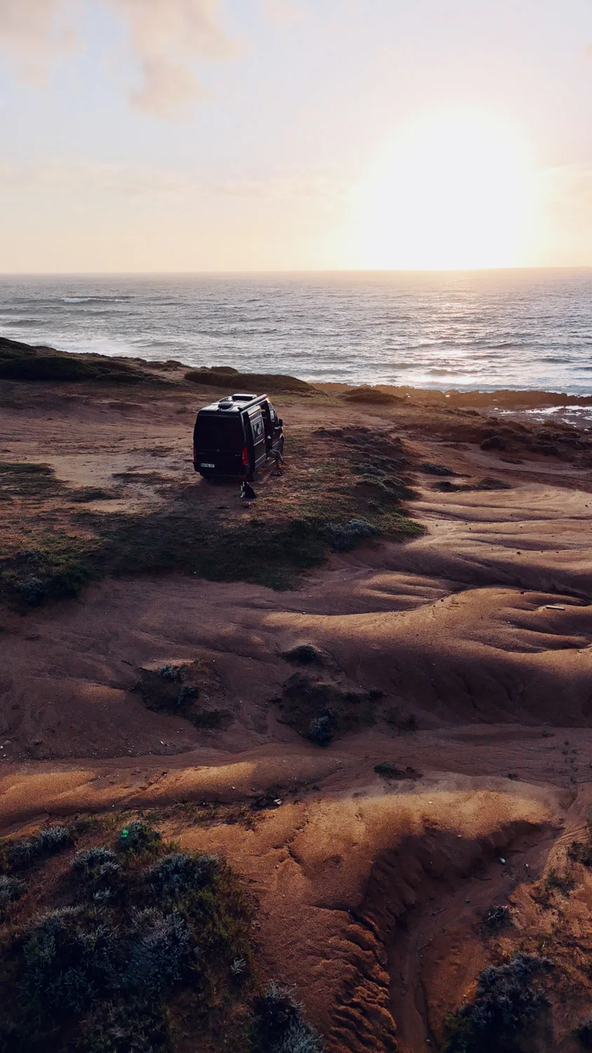 A black camper van parked on a coastal cliff at sunset with a person and a dog nearby overlooking the ocean.