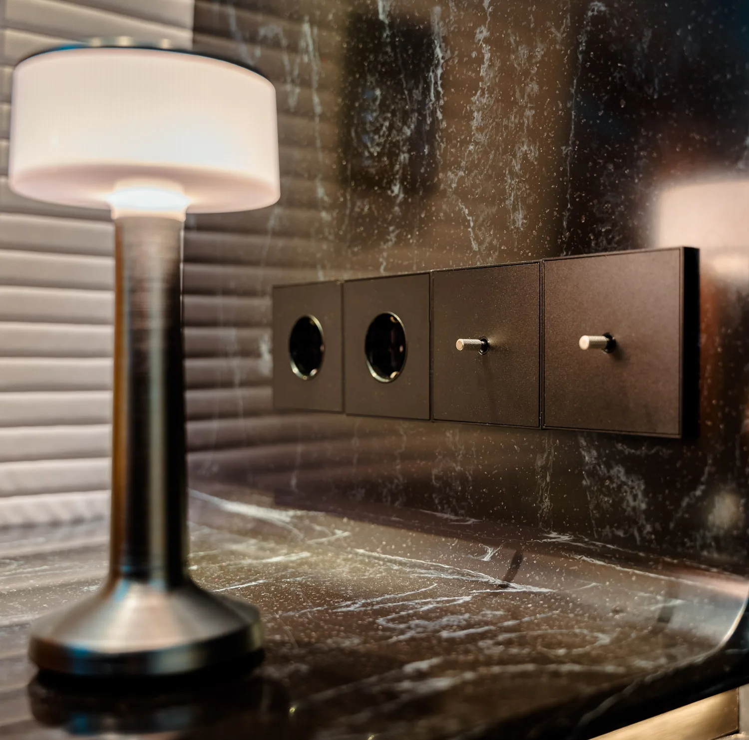 Black table lamp with a white lampshade on a marble surface near four dark square wall switches and sockets.