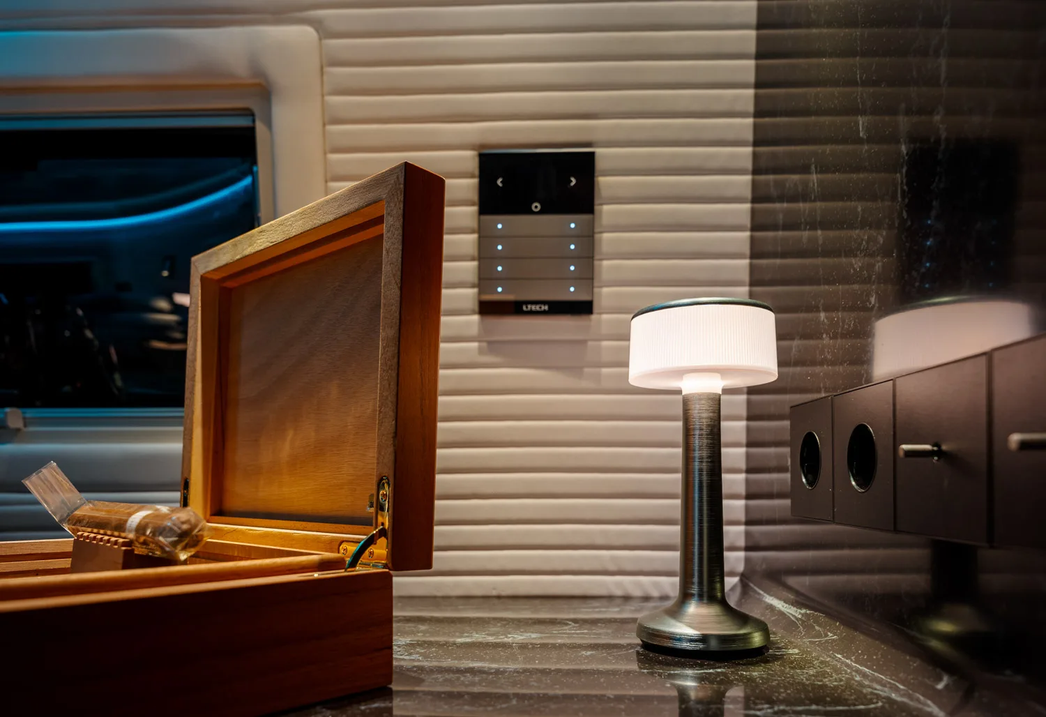 Open wooden cigar box with a wrapped cigar inside, next to a modern small table lamp on a marble surface with a textured beige wall and a control panel in the background.