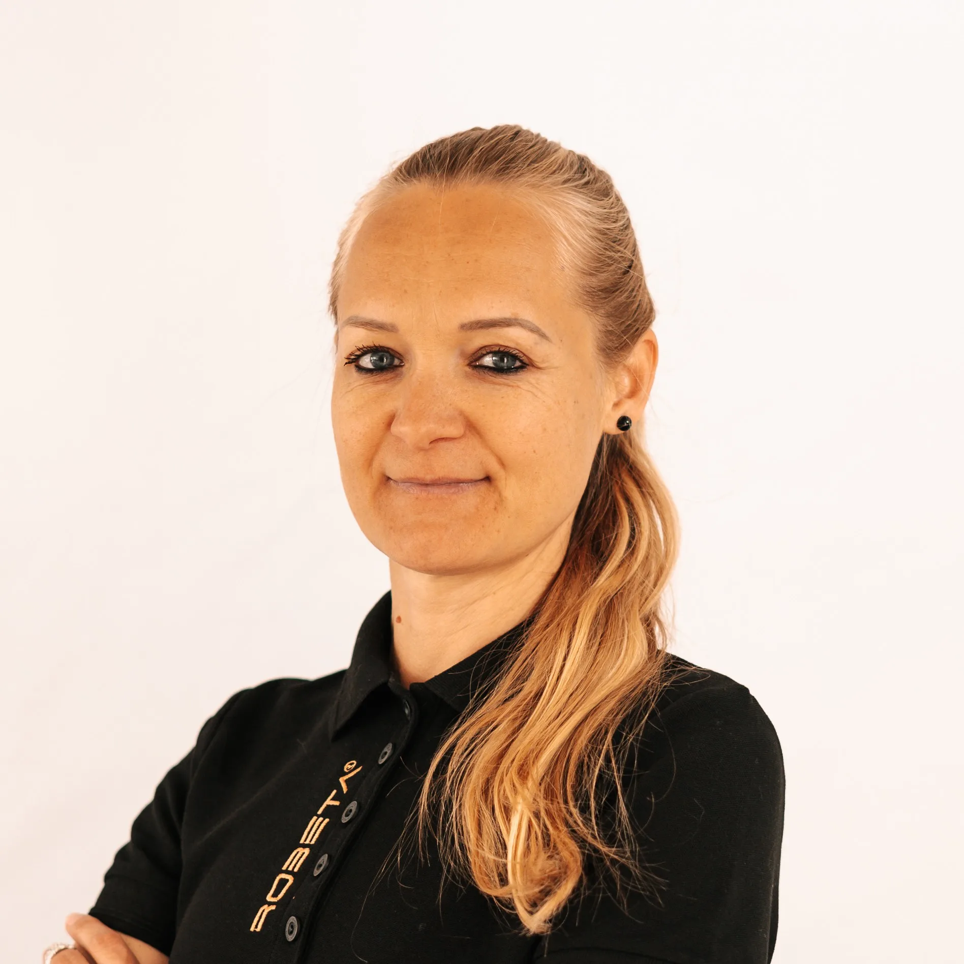 Smiling woman with long blonde hair in a black polo shirt with crossed arms against a plain white background.